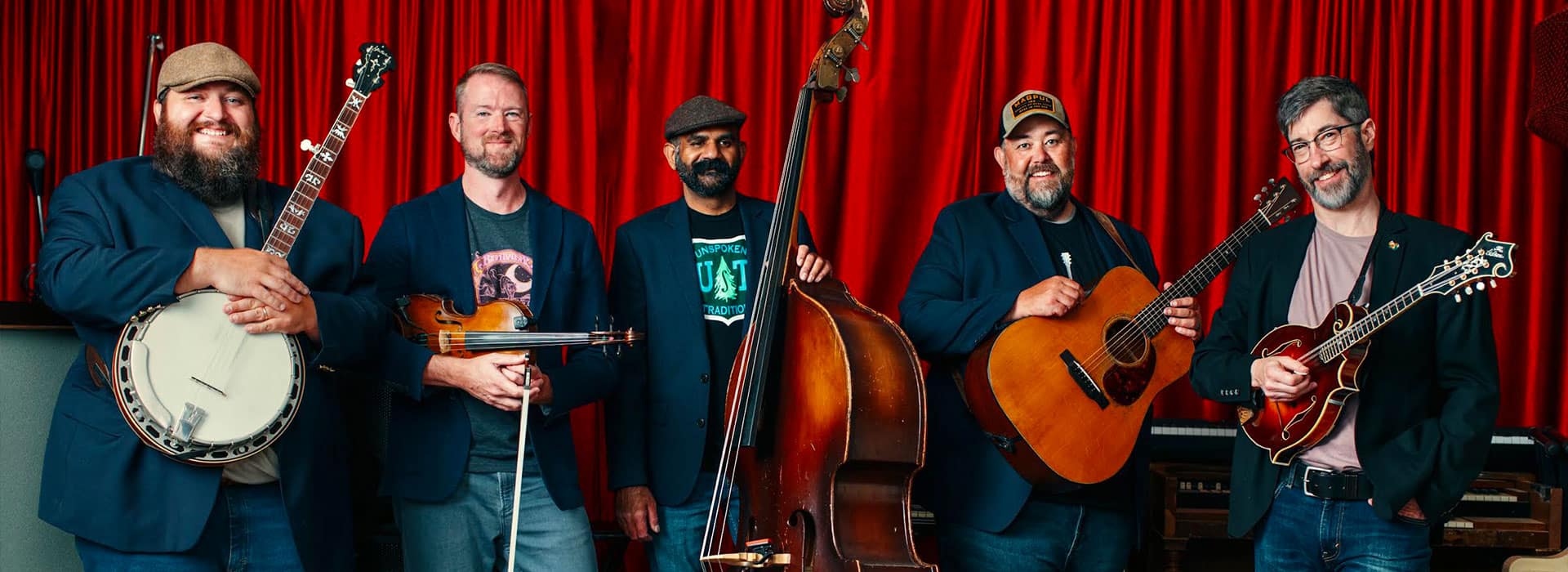 Five male musicians in blue jackets pose with their instruments&mdash;a banjo, violin, upright bass, acoustic guitar, and mandolin&mdash;against a red stage curtain.